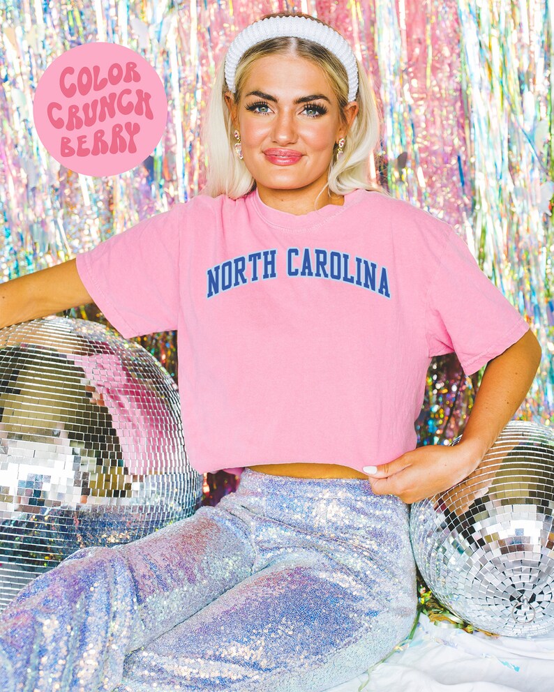 May include: A pink t-shirt with "NORTH CAROLINA" printed in navy blue. The image also features a white headband, sequined pants, and a disco ball. The text "COLOR CRUNCH BERRY" is in a pink circle.