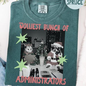 May include: A blue spruce coloured t-shirt featuring a black and white graphic of an opossum, a raccoon, and a small tree dressed in holiday attire at an office desk. The text reads JOLLIEST BUNCH OF ADMINISTRATORS with light green star accents.
