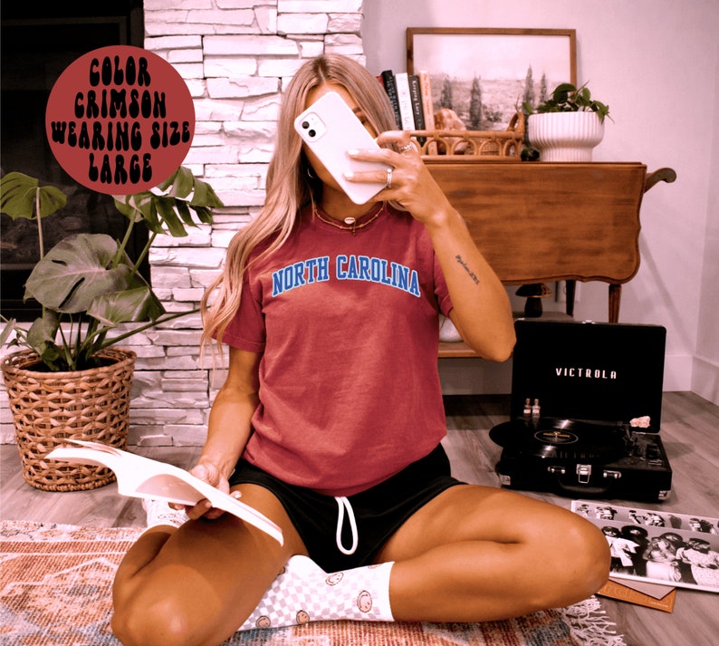 May include: Crimson t-shirt with "North Carolina" in blue arched text. The person is wearing black shorts and white socks with a smiley face pattern. A black Victrola record player is in the background.