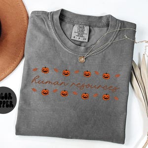 May include: A gray Comfort Colors t-shirt with the words "human resources" in a brown script font. The shirt features a row of orange jack-o'-lanterns and leaves. A gold necklace with a pendant is on the shirt.