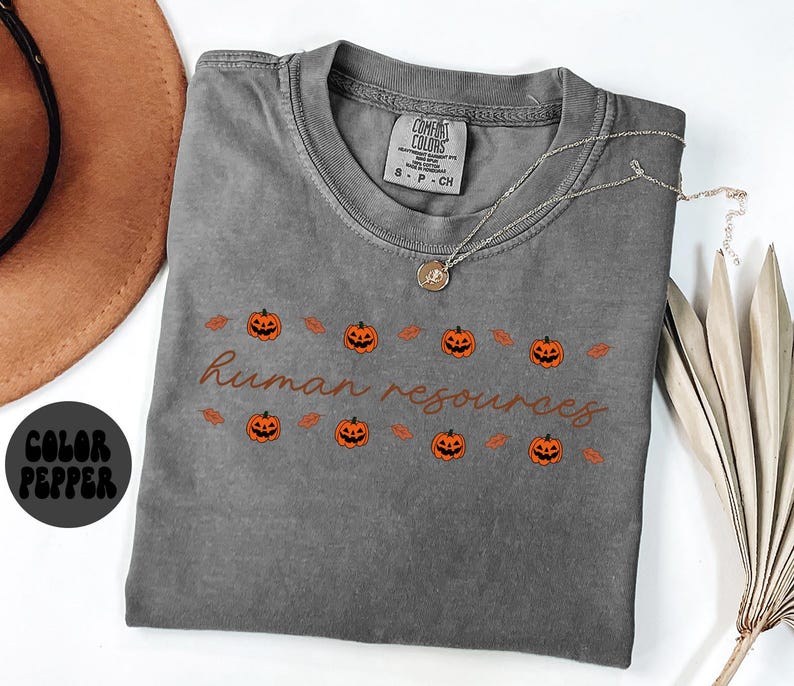 May include: A gray Comfort Colors t-shirt with the words "human resources" in a brown script font. The shirt features a row of orange jack-o'-lanterns and leaves. A gold necklace with a pendant is on the shirt.