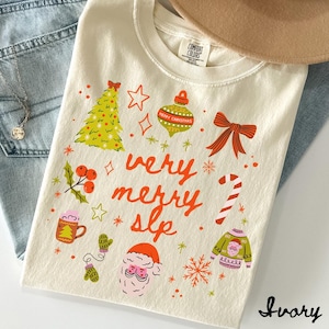 Peut inclure: T-shirt ivoire avec un motif festif. Le t-shirt présente un sapin de Noël, des ornements et les mots "very merry slp" en orange. D'autres graphiques incluent un visage de Père Noël, des flocons de neige et un sucre d'orge.