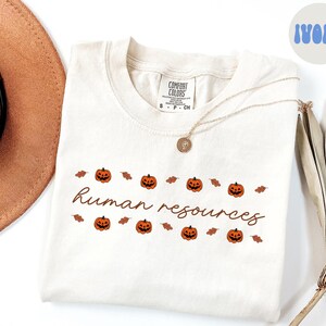 May include: Ivory-colored t-shirt with the words "human resources" in a cursive font, decorated with orange jack-o'-lanterns and brown leaves. A gold necklace and a brown hat are also visible.