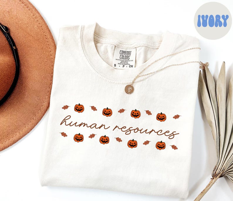 May include: Ivory-colored t-shirt with the words "human resources" in a cursive font, decorated with orange jack-o'-lanterns and brown leaves. A gold necklace and a brown hat are also visible.
