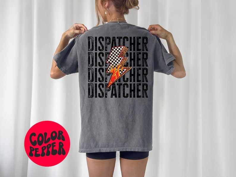 以下が含まれることがあります： グレーのTシャツに、黒い文字で「DISPATCHER」という単語が4回繰り返されています。オレンジ色の炎がついた黒と白の市松模様の稲妻がTシャツの中央に配置されています。