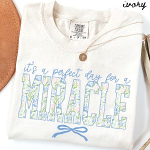 Puede incluir: Camiseta de color marfil con la frase "it's a perfect day for a MIRACLE" en letras azules y bloques florales. Un lazo azul está debajo de la palabra "MIRACLE". Un collar dorado con un colgante está sobre la camiseta.