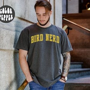 K&ouml;nnte beinhalten: Ein dunkelgraues T-Shirt mit dem Text "BIRD NERD" in gelben Buchstaben.