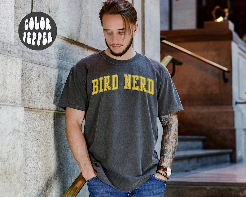 K&ouml;nnte beinhalten: Ein dunkelgraues T-Shirt mit dem Text "BIRD NERD" in gelben Buchstaben.