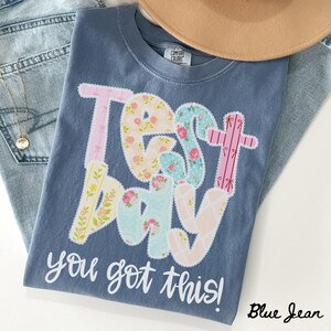 Könnte beinhalten: Blaues Comfort Colors T-Shirt mit den Worten "Test Day" in einem floralen Patchwork-Design. Der Satz "you got this!" ist darunter gedruckt. Ein beiger Hut und blaue Jeans sind im Hintergrund.
