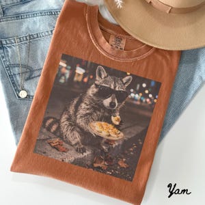 May include: A rust-colored crew neck t-shirt featuring a graphic print of a raccoon wearing black sunglasses and eating deviled eggs on a plate. The shirt has a Comfort Colors label at the neck and is displayed with blue denim and a tan hat.