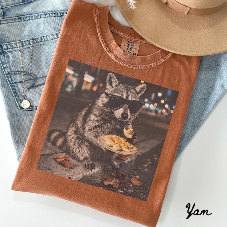 May include: A rust-colored crew neck t-shirt featuring a graphic print of a raccoon wearing black sunglasses and eating deviled eggs on a plate. The shirt has a Comfort Colors label at the neck and is displayed with blue denim and a tan hat.