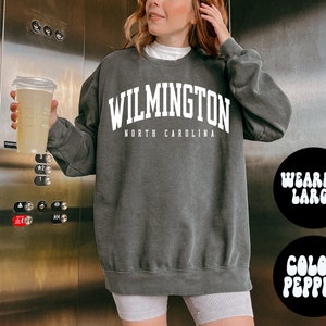 May include: A dark grey sweatshirt with the text "WILMINGTON NORTH CAROLINA" in white. The model is wearing a large size.