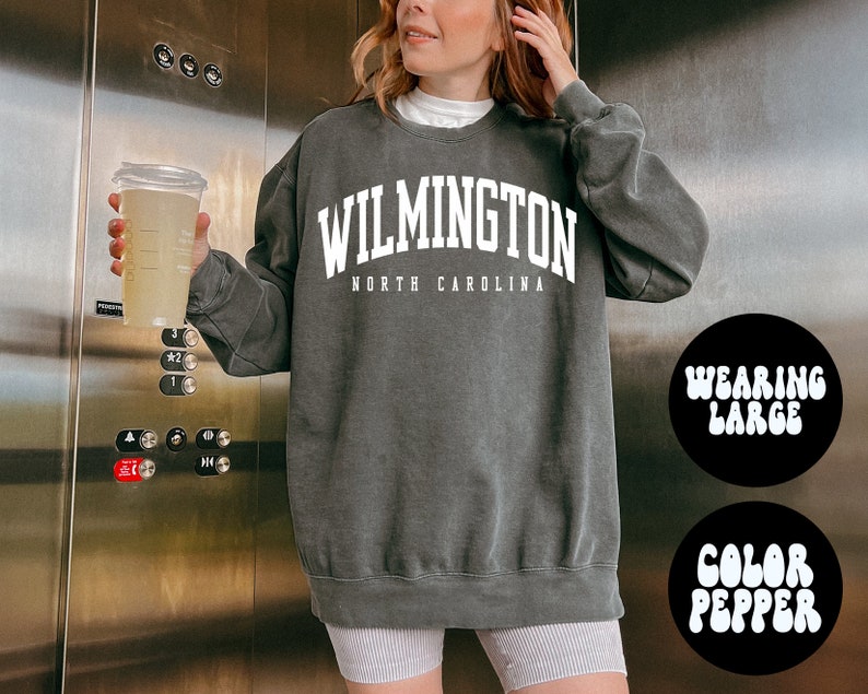 May include: A dark grey sweatshirt with the text "WILMINGTON NORTH CAROLINA" in white. The model is wearing a large size.