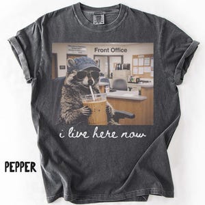 May include: A charcoal grey t-shirt featuring a graphic of a raccoon wearing a blue baseball cap and sunglasses, sitting at a desk in an office labelled Front Office, holding an iced coffee with a straw, with the text i live here now below.