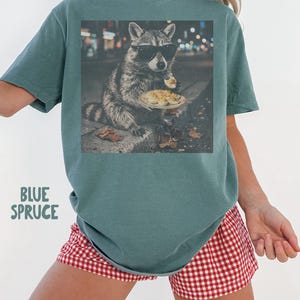May include: A blue spruce colored t-shirt featuring a graphic print of a raccoon wearing black sunglasses and eating deviled eggs on a plate. The text "BLUE SPRUCE" is printed in a light green font on the lower left side of the shirt.