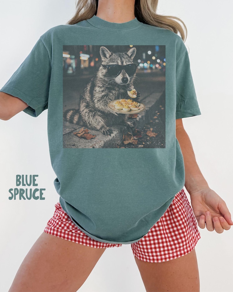 May include: A blue spruce colored t-shirt featuring a graphic print of a raccoon wearing black sunglasses and eating deviled eggs on a plate. The text "BLUE SPRUCE" is printed in a light green font on the lower left side of the shirt.