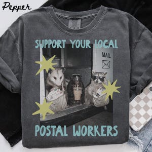 Könnte beinhalten: Ein anthrazitfarbenes T-Shirt mit einer Grafik, die ein Opossum, ein Stinktier und einen Waschbären mit Sonnenbrille zeigt, die Post halten. Der Text "SUPPORT YOUR LOCAL POSTAL WORKERS" ist in Hellblau gehalten und mit gelben Sternen verziert.