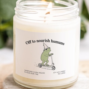 A white candle with a label featuring a frog riding a scooter, accompanied by the text "Off to nourish humans".