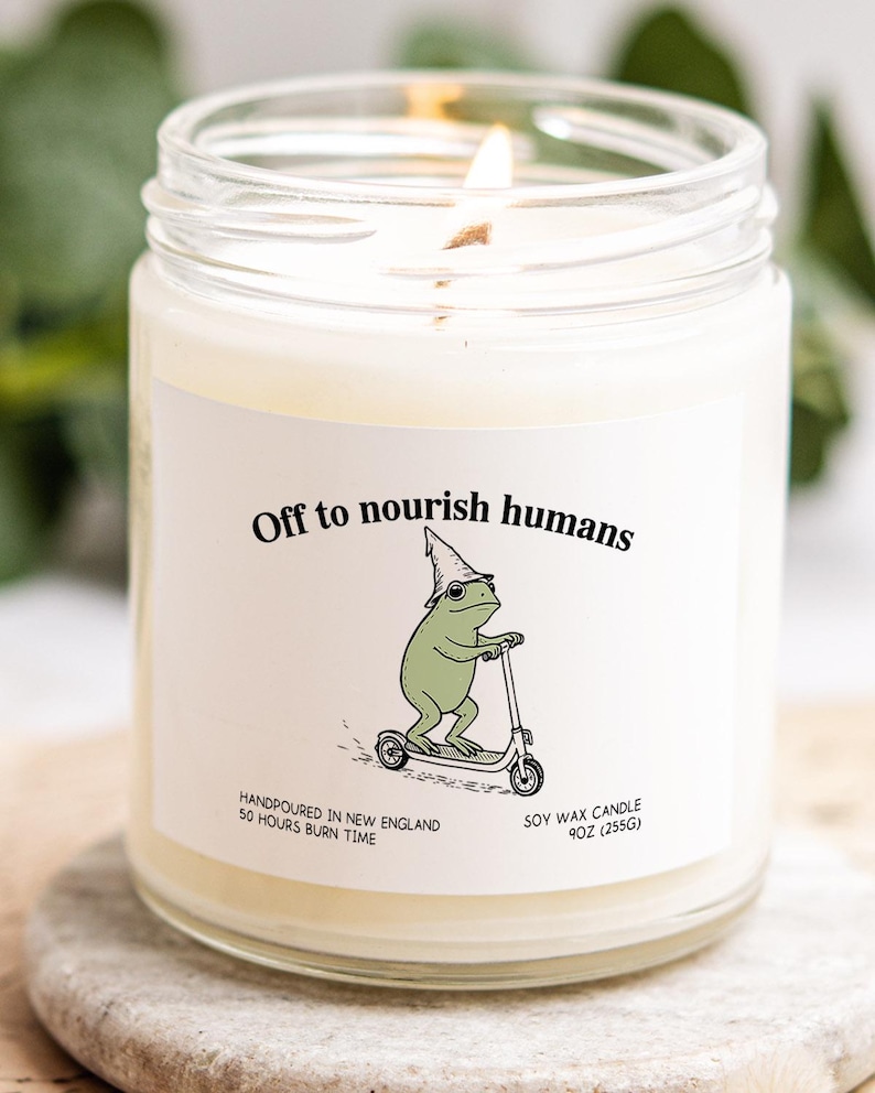 A white candle with a label featuring a frog riding a scooter, accompanied by the text "Off to nourish humans".