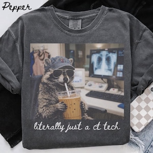 May include: A charcoal gray t-shirt featuring a graphic of a raccoon wearing a baseball cap and sunglasses, holding a plastic cup with a straw. The raccoon is positioned in front of a computer monitor displaying an X-ray. Text reads: literally just a ct tech.