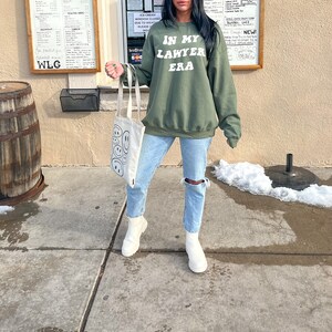 May include: A green sweatshirt with the text "IN MY LAWYER ERA" in white lettering. The person wearing the sweatshirt is holding a white tote bag with black smiley faces.