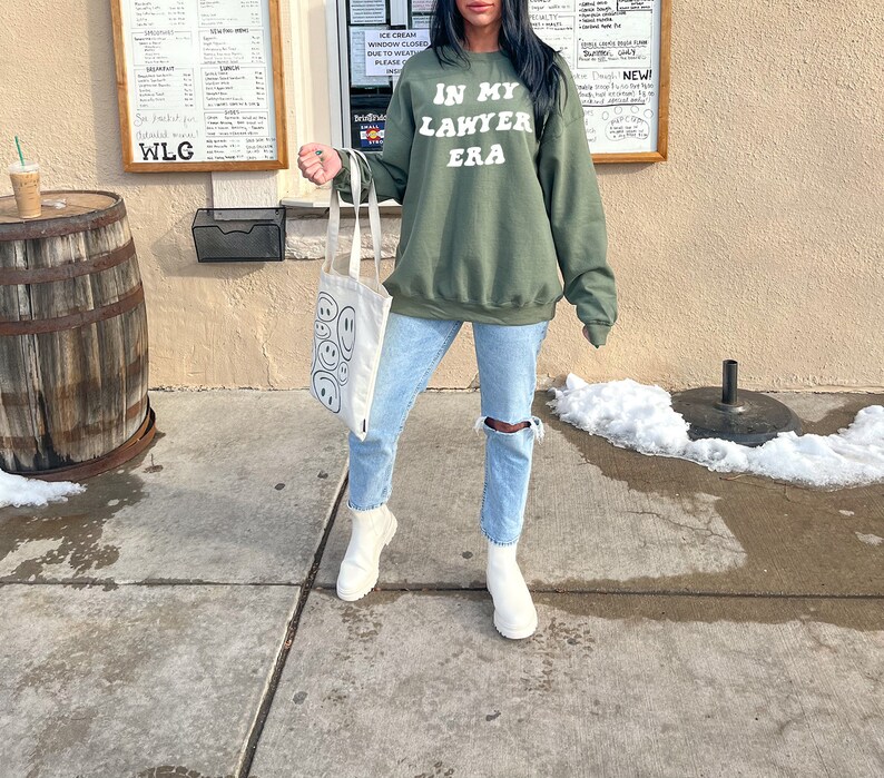 May include: A green sweatshirt with the text "IN MY LAWYER ERA" in white lettering. The person wearing the sweatshirt is holding a white tote bag with black smiley faces.