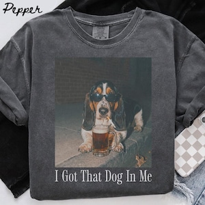 Può includere: Una t-shirt a girocollo grigio antracite con una stampa grafica di un basset hound che indossa occhiali da sole neri e tiene una sigaretta in bocca accanto a un boccale di birra. Il testo "I Got That Dog In Me" è stampato in bianco sotto l'immagine.