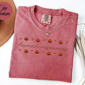 May include: Crimson-colored t-shirt with the text "human resources" and pumpkin graphics. The shirt has a crew neck and short sleeves. A silver necklace with a pendant is draped across the shirt.
