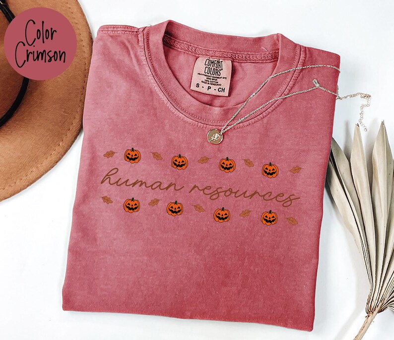 May include: Crimson-colored t-shirt with the text "human resources" and pumpkin graphics. The shirt has a crew neck and short sleeves. A silver necklace with a pendant is draped across the shirt.