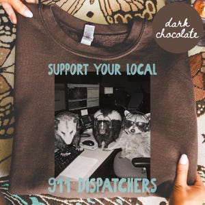 May include: A dark chocolate brown crewneck sweatshirt featuring a graphic print of an opossum, a skunk, and a raccoon wearing headsets at a computer desk. The text above reads SUPPORT YOUR LOCAL and the text below reads 911 DISPATCHERS.