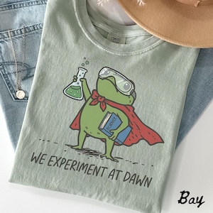Może przedstawiać: Szałwiowo-zielony t-shirt z grafiką zielonej żaby w goglach ochronnych i czerwonej pelerynie. Żaba trzyma zlewkę z zielonym płynem oraz niebieską książkę z napisem SCIENCE. Poniżej znajduje się czarny napis o treści WE EXPERIMENT AT DAWN.
