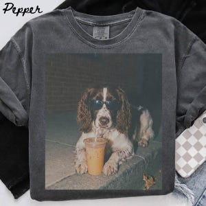Może przedstawiać: Ciemnoszary t-shirt z okrągłym dekoltem z nadrukiem przedstawiającym brązowo-białego spaniela w czarnych okularach przeciwsłonecznych, pijącego z przezroczystego plastikowego kubka ze słomką. Koszulka ma wykończenie w stylu vintage z efektem sprania.