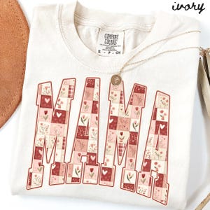 May include: Ivory-colored Comfort Colors t-shirt with the word "MAMA" in a patchwork design of red, pink, and white floral patterns. A gold necklace with a pendant is draped across the shirt.