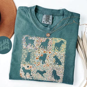 Op de afbeelding: Een teal Comfort Colors t-shirt met een bloemen- en honden silhouet ontwerp. Het shirt heeft een ronde hals en korte mouwen. Een gouden ketting met een hanger is over het shirt gedrapeerd. De tekst "BLUE SPRUCE" staat op een teal cirkel.