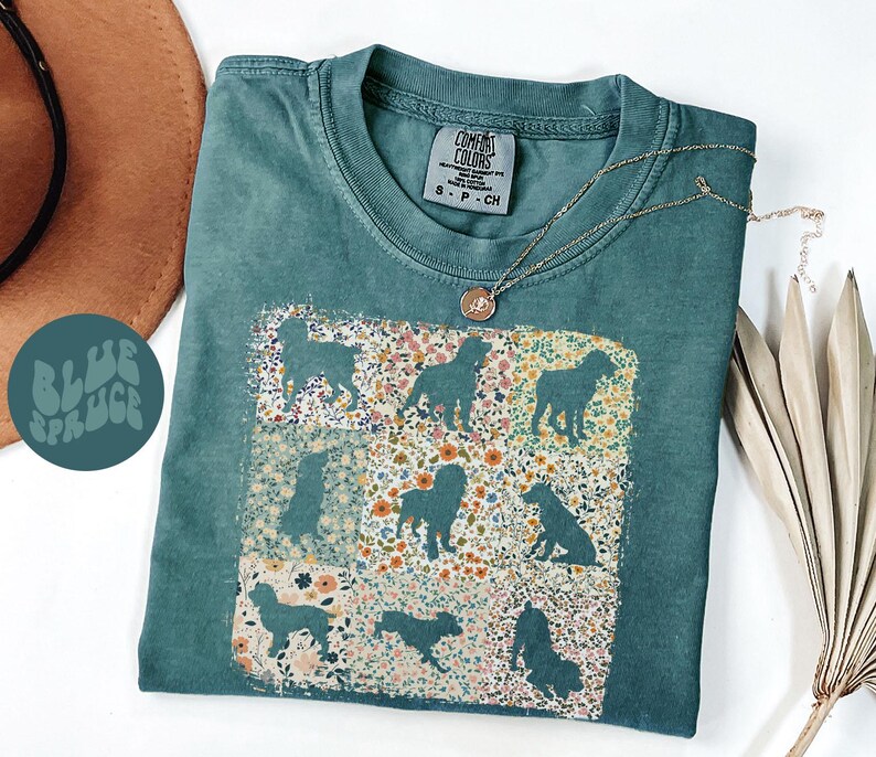 Op de afbeelding: Een teal Comfort Colors t-shirt met een bloemen- en honden silhouet ontwerp. Het shirt heeft een ronde hals en korte mouwen. Een gouden ketting met een hanger is over het shirt gedrapeerd. De tekst "BLUE SPRUCE" staat op een teal cirkel.