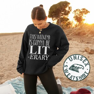 May include: Black sweatshirt with white text that reads "This weekend is gonna be LIT -ERARY". The sweatshirt is being worn by a person kneeling on a blanket with a pizza box and a camera in the foreground. The image also includes a green and white unisex sweatshirt logo.