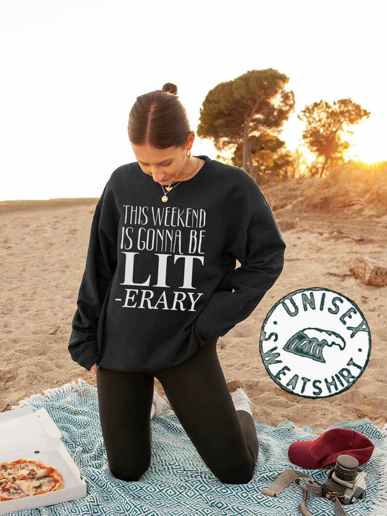 May include: Black sweatshirt with white text that reads "This weekend is gonna be LIT -ERARY". The sweatshirt is being worn by a person kneeling on a blanket with a pizza box and a camera in the foreground. The image also includes a green and white unisex sweatshirt logo.
