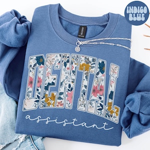 Op de afbeelding: Een blauwe sweatshirt met de woorden "DENTAL assistant" in een bloemenpatroon. De sweatshirt heeft een ronde hals en lange mouwen. Een zilveren ketting met een zonnehanger is over de voorkant gedrapeerd. De woorden "Indigo Blue" staan in een cirkel rechtsboven.