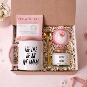 Könnte beinhalten: Geschenkbox mit einer weißen Tasse mit rosa Griff, einem Teebeutel, einem goldenen Löffel, einer Badekugel und einer Kerze. Auf der Tasse steht "THE LIFE OF AN IVF MAMA". Die Kerze ist mit "WHITE SAGE + LAVENDAR" beschriftet.