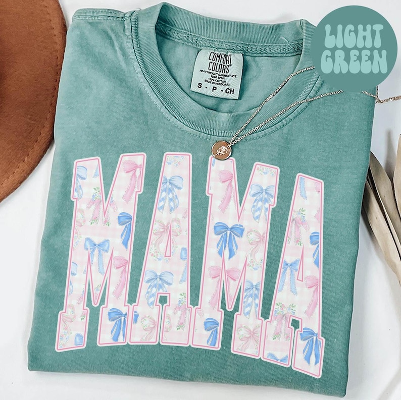 A green t-shirt with the word "MAMA" printed in pink and blue floral designs, along with a necklace and a brown hat.