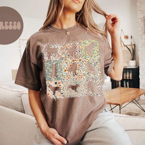 Op de afbeelding: Espresso-kleurig T-shirt met een bloemenontwerp. Het shirt heeft een rechthoekige afbeelding gevuld met een mix van kleurrijke bloemen en bladeren. Het woord "Espresso" is gedrukt in een cirkelvormige bruine vorm in de linkerbovenhoek. Het shirt heeft korte mouwen en een relaxte pasvorm.