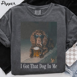 May include: A charcoal grey crew neck t-shirt featuring a graphic of a brown spaniel dog wearing black sunglasses, holding a cigarette in its mouth, and resting its paws next to a glass of beer. The text "I Got That Dog In Me" is printed below.