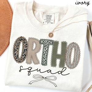 Op de afbeelding: Ivoor kleurig T-shirt met de woorden "ORTHO squad" in een decoratief lettertype. De letters hebben verschillende patronen, waaronder camouflage, luipaardprint en strepen. Een gouden ketting met een hanger ligt over het shirt.