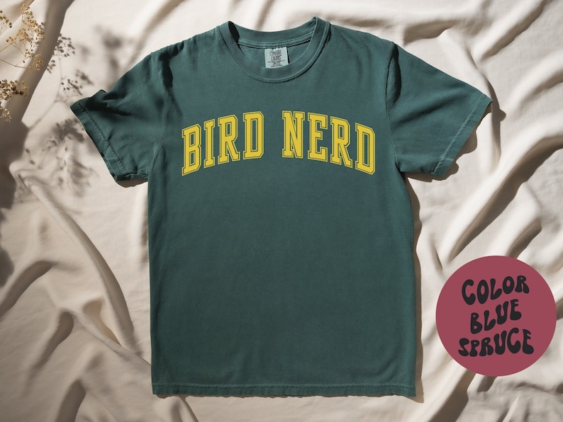 K&ouml;nnte beinhalten: Ein dunkelgr&uuml;nes T-Shirt mit dem Text "BIRD NERD" in gelben Buchstaben. Das Shirt hat einen Vintage-Look und -Feel.
