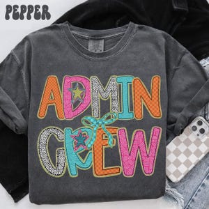 Op de afbeelding: Donkergrijs sweatshirt met de woorden "ADMIN CREW" in kleurrijke, patroonletters. Het ontwerp bevat sterren en een strik. Het woord "PEPPER" staat in de linkerbovenhoek van de afbeelding.