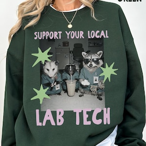 May include: A forest green crewneck sweatshirt featuring a graphic print of an opossum, a skunk, and a raccoon dressed in teal scrubs. The text reads SUPPORT YOUR LOCAL LAB TECH in light pink letters, accented by light green star shapes.