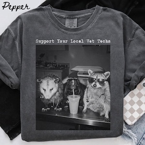 May include: A charcoal gray crew neck t-shirt featuring a black and white graphic print of an opossum, a skunk, and a raccoon wearing stethoscopes. The text above the graphic reads Support Your Local Vet Techs. The raccoon is wearing sunglasses.