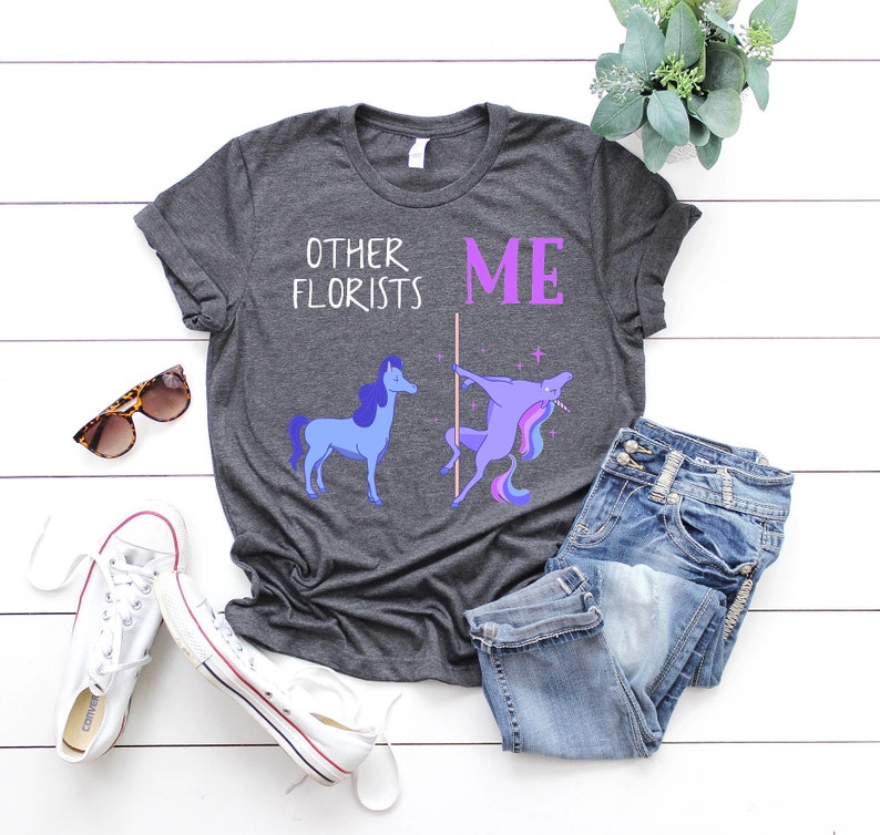 May include: A dark gray t-shirt with a humorous graphic of two unicorns. The text on the shirt reads "Other Florists" and "Me". The unicorn on the left is standing normally, while the unicorn on the right is doing a pole dance.