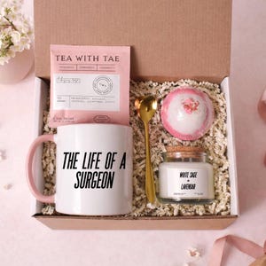 May include: A gift box containing a white mug with pink handle and the text "THE LIFE OF A SURGEON", a pink tea packet, a gold spoon, a bath bomb, and a jar of white sage and lavender candle.