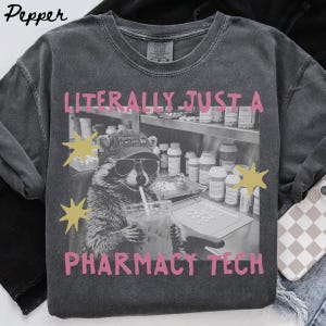 May include: A charcoal grey crew neck t-shirt featuring a black and white graphic of a raccoon wearing a cap that says Pharmacy is my jam, holding a drink with a straw. The text LITERALLY JUST A PHARMACY TECH is printed in pink with yellow stars.
